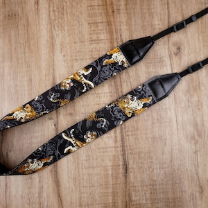 Puede incluir: Correa de cámara negra con diseño de estampado de tigre. La correa es ajustable y tiene una sección acolchada para el hombro.