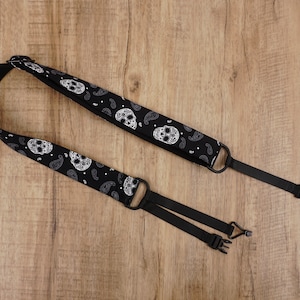 Może przedstawiać: Czarny pasek do aparatu z białym wzorem czaszki i paisley. Pasek jest regulowany i posiada metalowe i plastikowe okucia.