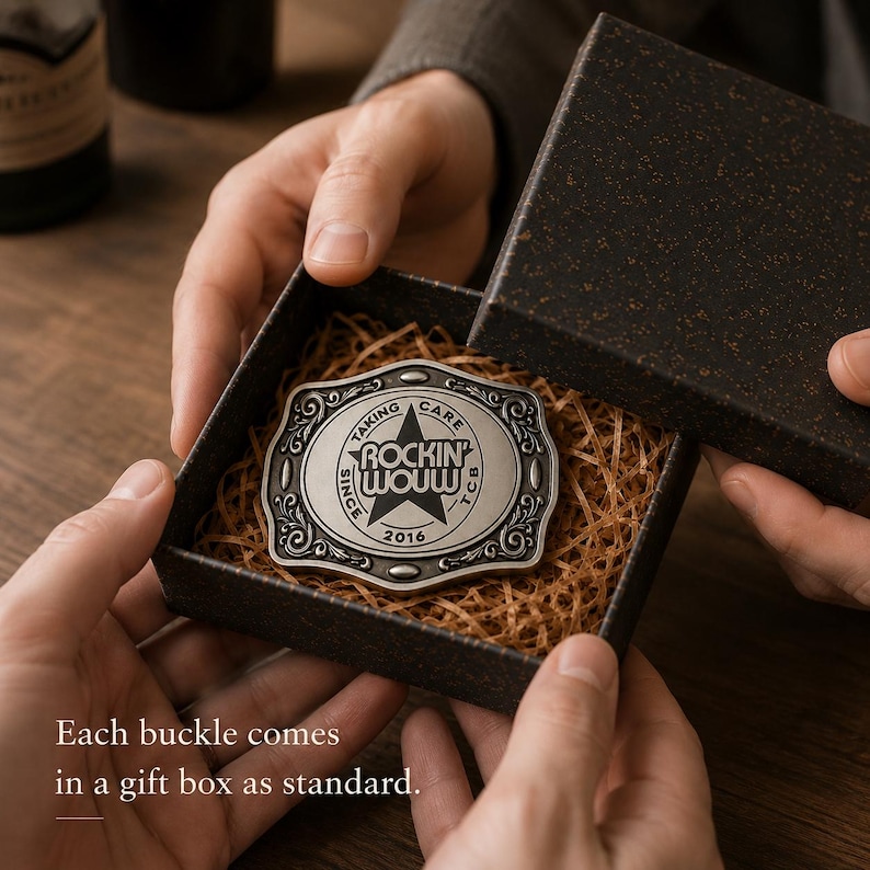 May include: A rectangular silver-toned metal belt buckle with ornate scrollwork edges and a central star design. The text reads ROCKIN' WOWW, TAKING CARE, TCB, and 2016. The buckle is nestled inside a dark textured gift box filled with shredded paper.