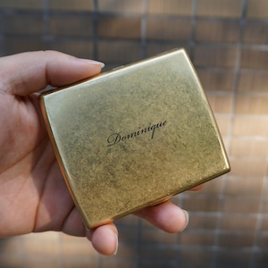 May include: A rectangular, gold-toned metal compact case with a weathered, textured finish. The name Dominique is engraved in a dark, elegant script font in the centre of the lid. The compact has a slim, flat profile with rounded corners.