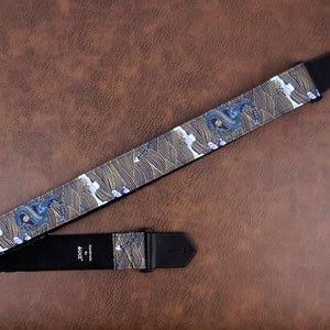 Op de afbeelding: Een zwarte gitaarband met een blauw en gouden draken- en golfpatroon. De band heeft een zwart leren uiteinde met het woord "AJH" erop gedrukt.