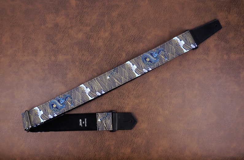 Op de afbeelding: Een zwarte gitaarband met een blauw en gouden draken- en golfpatroon. De band heeft een zwart leren uiteinde met het woord "AJH" erop gedrukt.