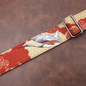 May include: A red and gold guitar strap with a Japanese crane design. The strap has a silver metal buckle.
