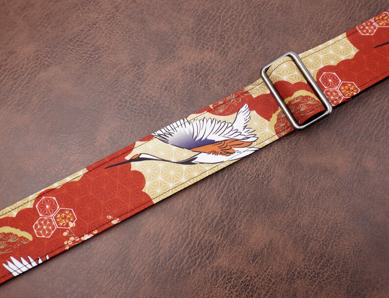 May include: A red and gold guitar strap with a Japanese crane design. The strap has a silver metal buckle.