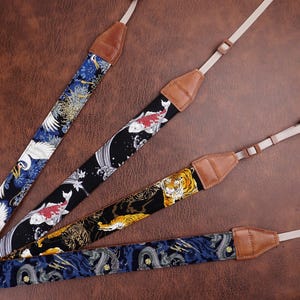 Könnte beinhalten: Vier Kameragurte mit japanisch inspirierten Designs. Die Gurte zeigen Muster von Kranichen, Koi-Karpfen, Tigern und Drachen in Blau, Schwarz und Gold. Jeder Gurt hat einen beigefarbenen Nackengurt und braune Lederenden.