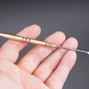 Może przedstawiać: Złote metalowe narzędzie z ostrym końcem i uchwytem grawerowanym literami "Y.W.B."