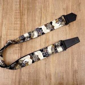 May include: A guitar strap featuring a black fabric base with a repeating pattern of white cranes and gold floral accents. The strap includes black leather end tabs with pre-cut holes and a metal adjustment buckle for length modification.
