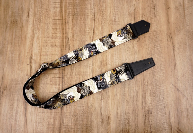 May include: A guitar strap featuring a black fabric base with a repeating pattern of white cranes and gold floral accents. The strap includes black leather end tabs with pre-cut holes and a metal adjustment buckle for length modification.