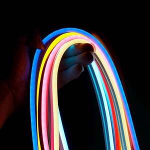 May include: A hand holding a flexible, multi-colored LED strip against a black background. The strip features vibrant colors including blue, yellow, pink, and red, creating a rainbow effect. The lighting is bright and the overall aesthetic is modern.
