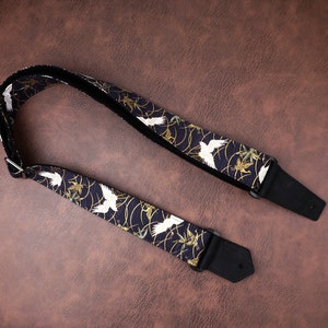 May include: A guitar strap featuring a dark navy blue fabric with a repeating pattern of white cranes and gold swirling lines. The strap includes black leather ends with pre-cut holes and a silver metal adjustment buckle for length modification.