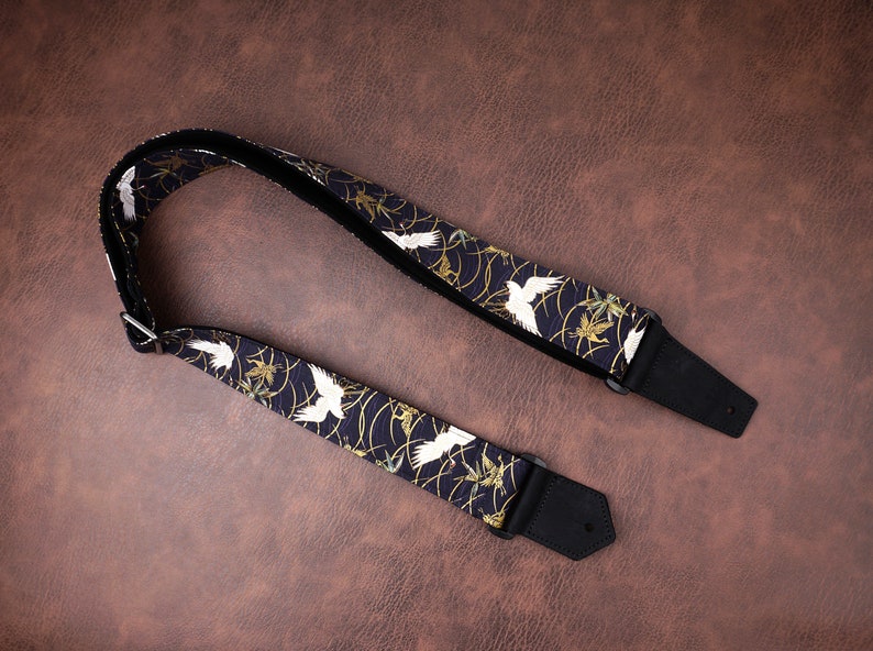 May include: A guitar strap featuring a dark navy blue fabric with a repeating pattern of white cranes and gold swirling lines. The strap includes black leather ends with pre-cut holes and a silver metal adjustment buckle for length modification.