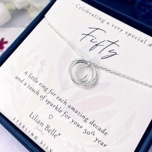 May include: A sterling silver necklace with three interlocking rings, one encrusted with small clear stones. The necklace is presented in a gift box with a card that reads "Fifty" and "A little ring for each amazing decade and a touch of sparkle for your 50th year."