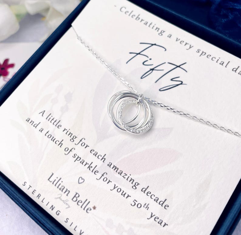 May include: A sterling silver necklace with three interlocking rings, one encrusted with small clear stones. The necklace is presented in a gift box with a card that reads "Fifty" and "A little ring for each amazing decade and a touch of sparkle for your 50th year."