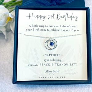 May include: A white card with the text "Happy 21st Birthday" and a silver necklace with a sapphire birthstone and two rings. The card says "A little ring to mark each decade and your birthstone to celebrate your 21st year." It also says "SAPPHIRE - symbolizing CALM, PEACE & TRANQUILITY." The card is signed "Lilian Belle Jewelry" and says "STERLING SILVER".