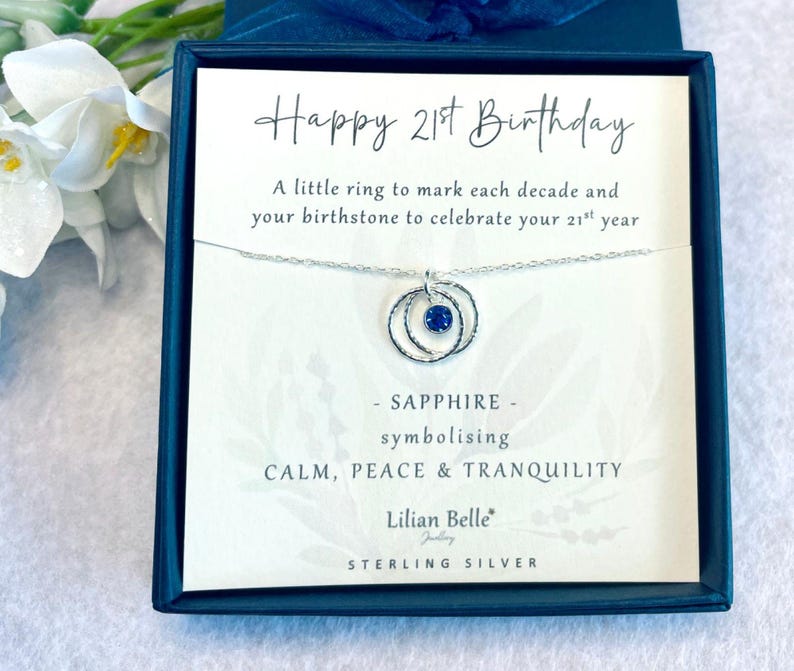 May include: A white card with the text "Happy 21st Birthday" and a silver necklace with a sapphire birthstone and two rings. The card says "A little ring to mark each decade and your birthstone to celebrate your 21st year." It also says "SAPPHIRE - symbolizing CALM, PEACE & TRANQUILITY." The card is signed "Lilian Belle Jewelry" and says "STERLING SILVER".