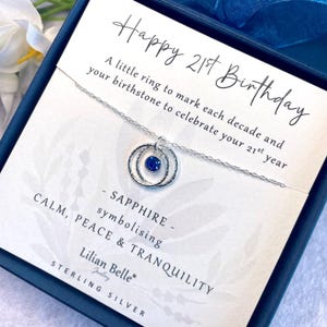 May include: A silver necklace with a sapphire birthstone pendant in a double ring design. The necklace comes in a box with the text "Happy 21st Birthday" and "A little ring to mark each decade and your birthstone to celebrate your 21st year."  The box also says "Sapphire - symbolizing Calm, Peace & Tranquility" and "Lilian Belle Sterling Silver."