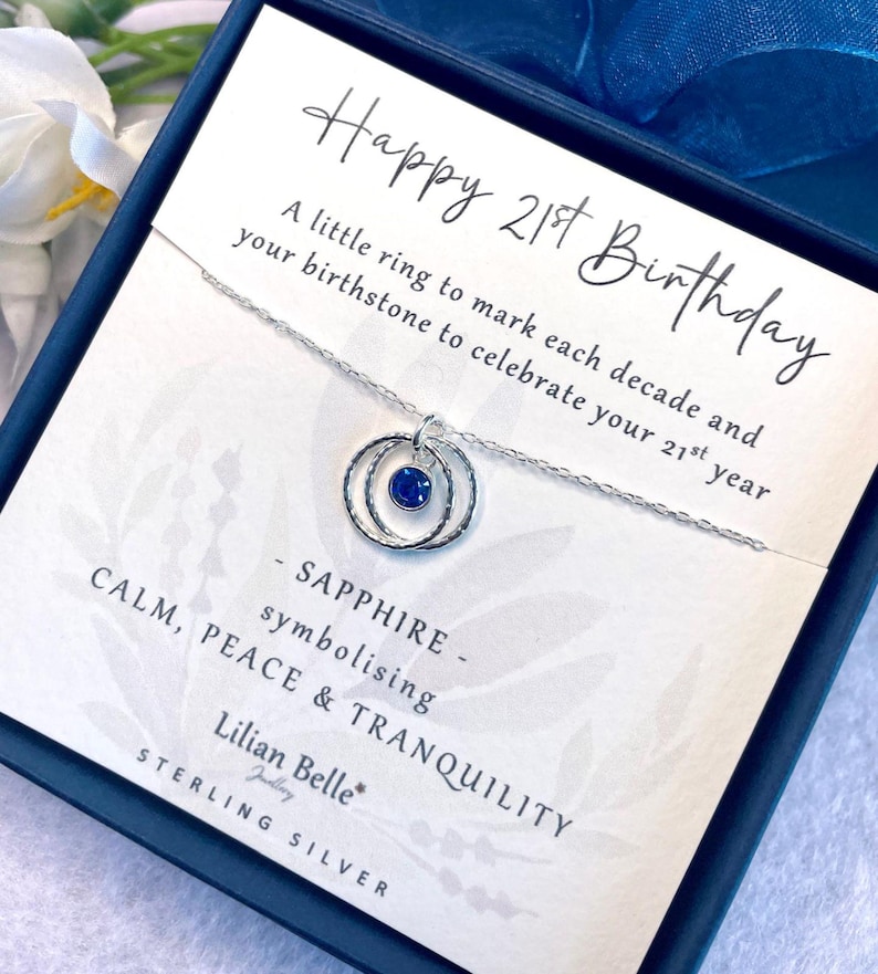 May include: A silver necklace with a sapphire birthstone pendant in a double ring design. The necklace comes in a box with the text "Happy 21st Birthday" and "A little ring to mark each decade and your birthstone to celebrate your 21st year."  The box also says "Sapphire - symbolizing Calm, Peace & Tranquility" and "Lilian Belle Sterling Silver."
