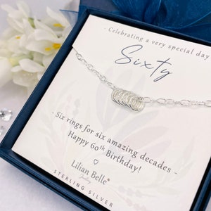 May include: A silver necklace with six small rings, presented in a blue box with a white card that reads "Celebrating a very special day Sixty Six rings for six amazing decades Happy 60th Birthday! Lilian Belle Jewelry STERLING SILVER".