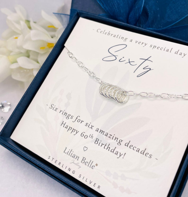 May include: A silver necklace with six small rings, presented in a blue box with a white card that reads "Celebrating a very special day Sixty Six rings for six amazing decades Happy 60th Birthday! Lilian Belle Jewelry STERLING SILVER".
