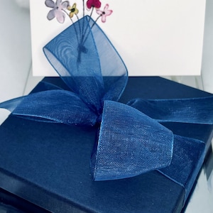 May include: A navy blue gift box with a blue sheer ribbon tied in a bow. The box is sitting on a white surface. The box has a white card on top with a hand-drawn bouquet of flowers in pink, yellow, and red.