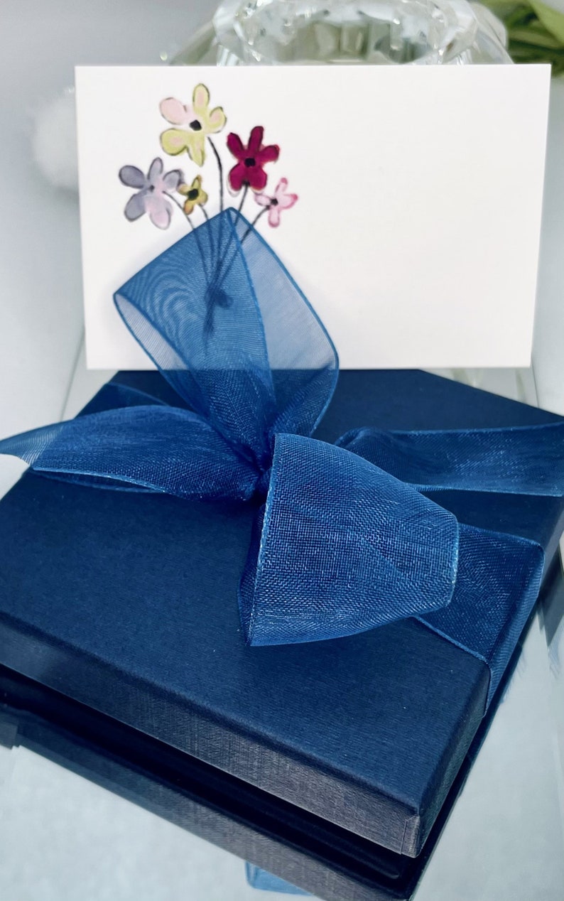 May include: A navy blue gift box with a blue sheer ribbon tied in a bow. The box is sitting on a white surface. The box has a white card on top with a hand-drawn bouquet of flowers in pink, yellow, and red.