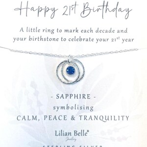May include: A silver necklace with three interlocking rings and a blue sapphire birthstone. The card reads "Happy 21st Birthday", "A little ring to mark each decade and your birthstone to celebrate your 21st year", "SAPPHIRE - symbolising CALM, PEACE & TRANQUILITY", "Lilian Belle Jewellery", "STERLING SILVER".