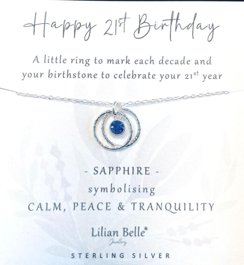 May include: A silver necklace with three interlocking rings and a blue sapphire birthstone. The card reads "Happy 21st Birthday", "A little ring to mark each decade and your birthstone to celebrate your 21st year", "SAPPHIRE - symbolising CALM, PEACE & TRANQUILITY", "Lilian Belle Jewellery", "STERLING SILVER".
