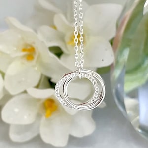 May include: A silver necklace with three interlocking rings, one ring is encrusted with small, clear gemstones. The necklace hangs from a delicate chain. The background features white flowers and a blurred glass object.