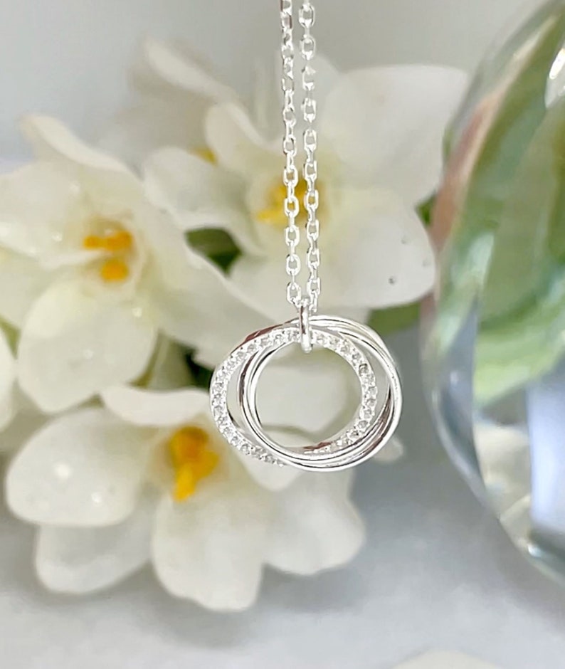 May include: A silver necklace with three interlocking rings, one ring is encrusted with small, clear gemstones. The necklace hangs from a delicate chain. The background features white flowers and a blurred glass object.