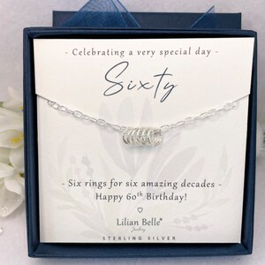 May include: A silver necklace with six rings, presented in a blue box with a white card that reads "Celebrating a very special day - Sixty - Six rings for six amazing decades - Happy 60th Birthday! - Lilian Belle Jewelry - STERLING SILVER".