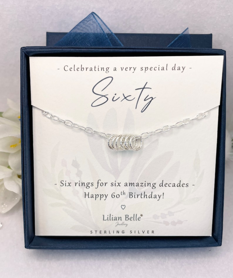 May include: A silver necklace with six rings, presented in a blue box with a white card that reads "Celebrating a very special day - Sixty - Six rings for six amazing decades - Happy 60th Birthday! - Lilian Belle Jewelry - STERLING SILVER".
