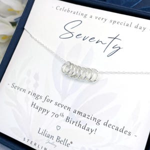 May include: A silver necklace with seven rings, packaged in a blue box with a card that reads "Celebrating a very special day Seventy - Seven rings for seven amazing decades - Happy 70th Birthday! Lilian Belle Jewellery STERLING SILVER".