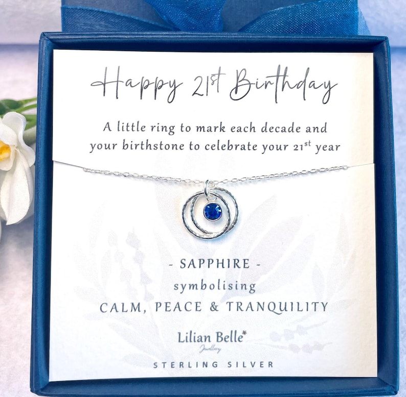 May include: A silver necklace with a blue sapphire birthstone and two rings, presented in a blue box with a card that reads "Happy 21st Birthday. A little ring to mark each decade and your birthstone to celebrate your 21st year. - SAPPHIRE - symbolising CALM, PEACE & TRANQUILITY. Lilian Belle Jewellery STERLING SILVER."