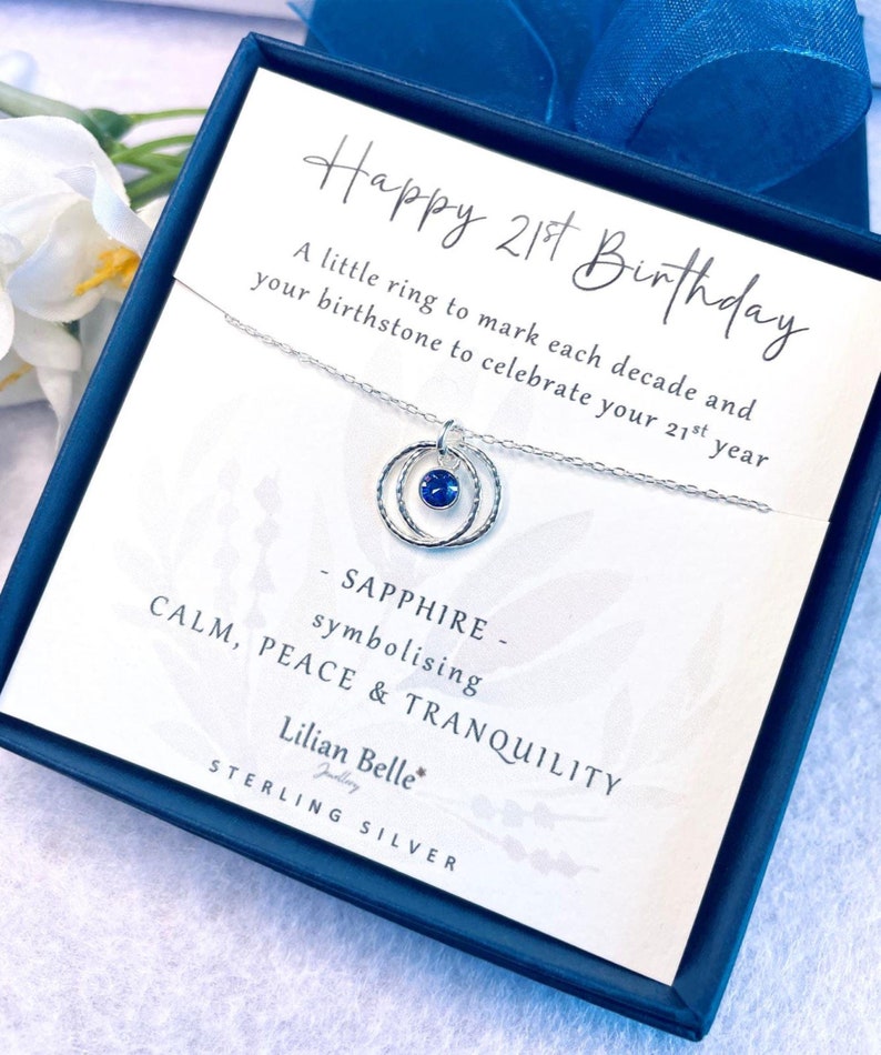 May include: A silver necklace with a blue sapphire gemstone pendant in a circle design. The necklace comes in a box with a card that says "Happy 21st Birthday" and "Sapphire - symbolizing Calm, Peace & Tranquility". Lilian Belle Jewelry Sterling Silver.