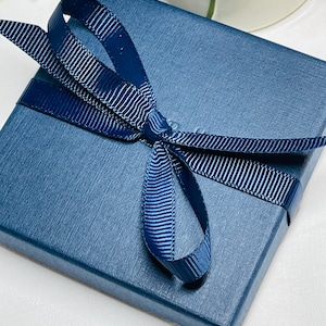May include: A navy blue gift box with a textured surface, tied with a matching ribbon and bow. The ribbon has a subtle striped pattern. The box is presented on a white surface, suggesting a gift or presentation setting.