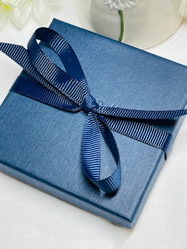 May include: A navy blue gift box with a textured surface, tied with a matching ribbon and bow. The ribbon has a subtle striped pattern. The box is presented on a white surface, suggesting a gift or presentation setting.