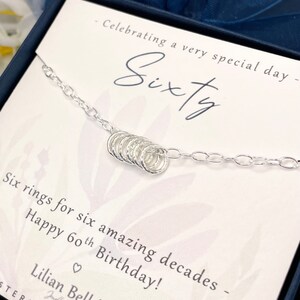 May include: A silver necklace with six small rings, presented in a box with the text "Celebrating a very special day - Sixty - Six rings for six amazing decades - Happy 60th Birthday! - Lilian Belle - Jewellery - STERLIN"