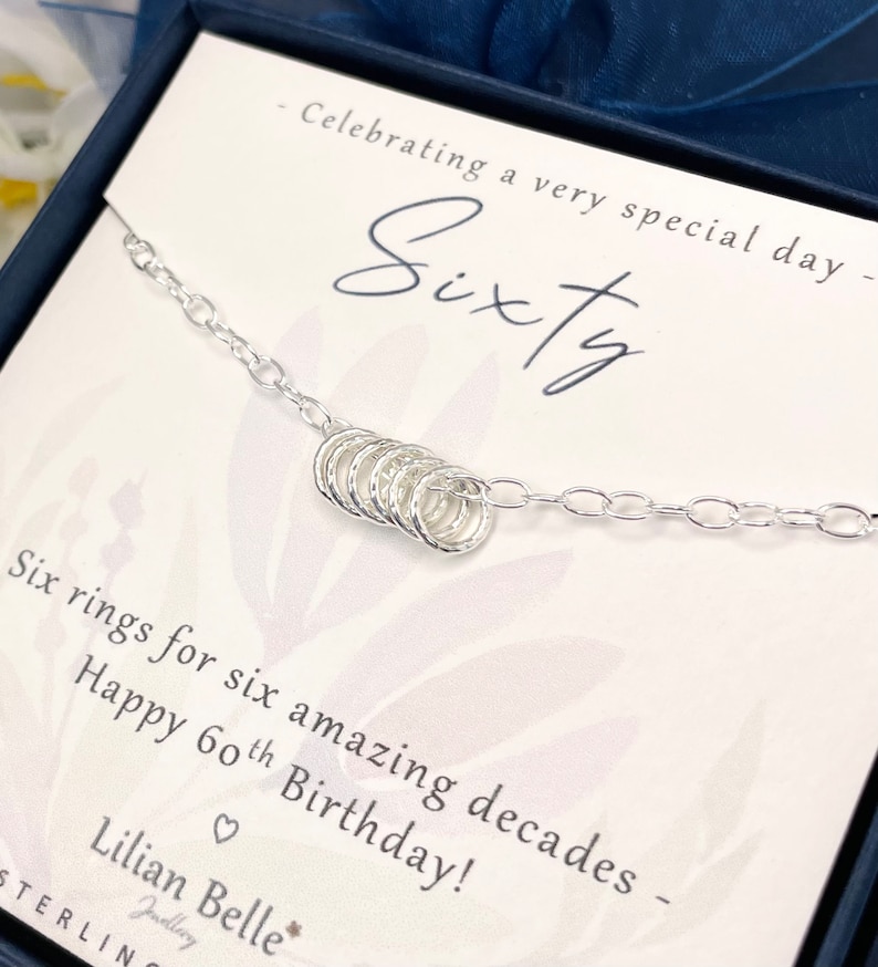 May include: A silver necklace with six small rings, presented in a box with the text "Celebrating a very special day - Sixty - Six rings for six amazing decades - Happy 60th Birthday! - Lilian Belle - Jewellery - STERLIN"