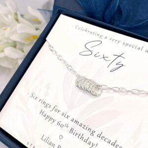 May include: A silver necklace with six small rings, a gift for a 60th birthday. The card reads "Celebrating a very special Sixty - Six rings for six amazing decades - Happy 60th Birthday! Lilian Belle Jewellery STERLING"