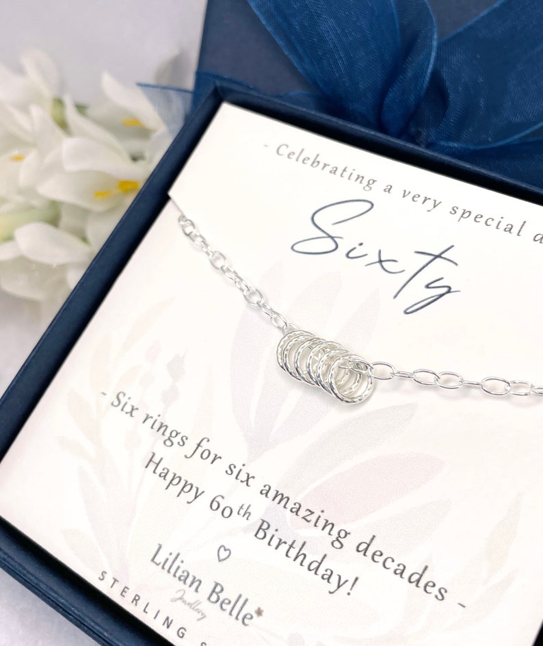 May include: A silver necklace with six small rings, a gift for a 60th birthday. The card reads "Celebrating a very special Sixty - Six rings for six amazing decades - Happy 60th Birthday! Lilian Belle Jewellery STERLING"