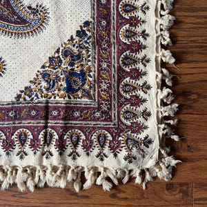 May include: Cream-colored textile with a paisley design in shades of blue, burgundy, and gold. The border features a repeating teardrop pattern. The edge is finished with cream-colored tassels. The textile is likely a decorative item, such as a tablecloth or wall hanging.