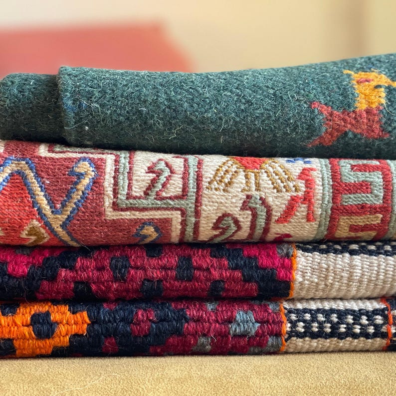 May include: A stack of four woven rugs in various colors and patterns. The top rug is a dark green, the second is a red and white patterned rug, the third is a red and black patterned rug, and the bottom rug is a white and black patterned rug.