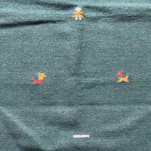 May include: A teal blue woven rug with a simple design of two yellow and red stylized figures and a white line across the bottom.