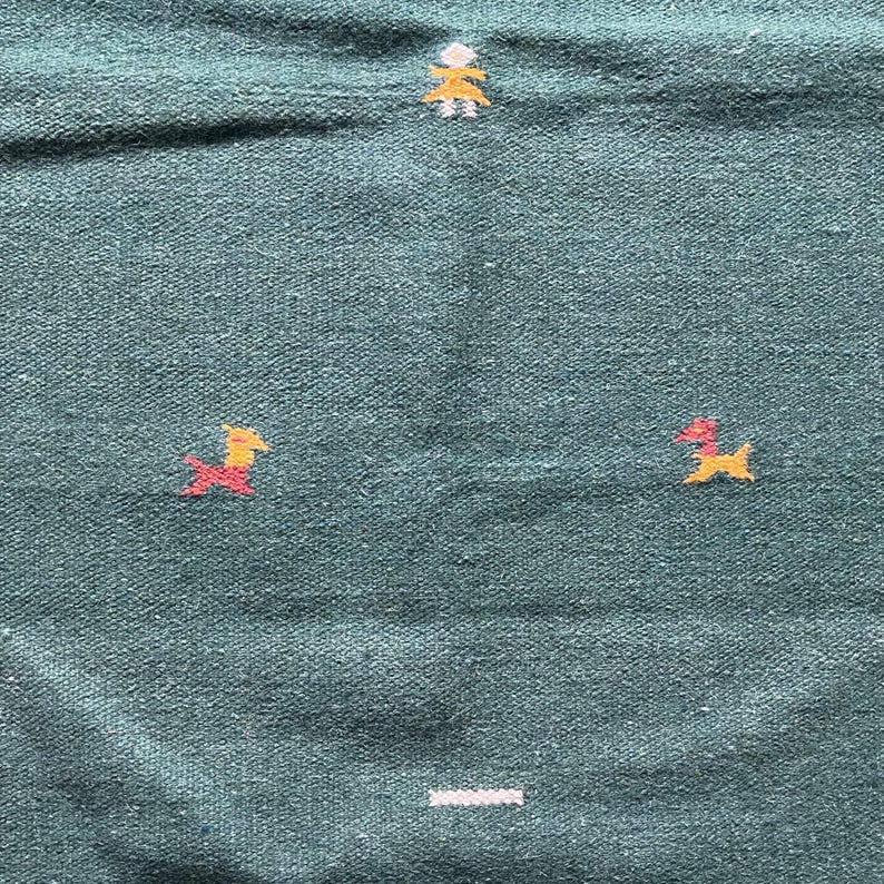 May include: A teal blue woven rug with a simple design of two yellow and red stylized figures and a white line across the bottom.