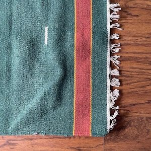 May include: A green woven rug with a red and yellow stripe down the center. The rug has white fringe on the edges.