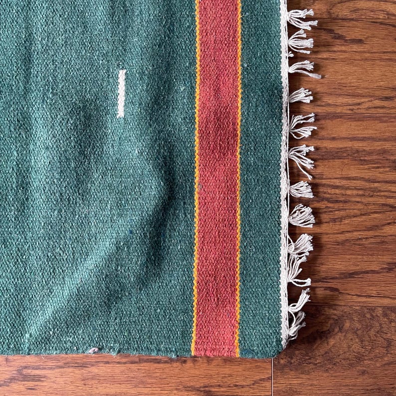 May include: A green woven rug with a red and yellow stripe down the center. The rug has white fringe on the edges.