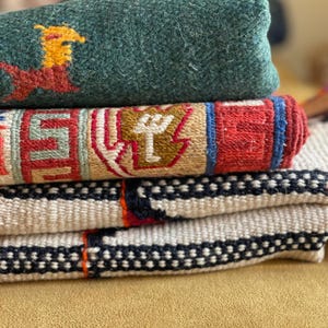 May include: A stack of four woven rugs in various patterns and colors. The top rug is a dark green wool rug with a red and yellow design. The second rug is a multi-colored rug with a geometric pattern. The third rug is a white rug with black and red stripes. The bottom rug is a white rug with black dots.