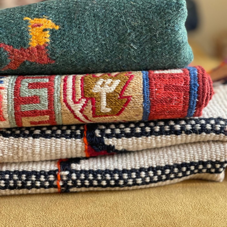 May include: A stack of four woven rugs in various patterns and colors. The top rug is a dark green wool rug with a red and yellow design. The second rug is a multi-colored rug with a geometric pattern. The third rug is a white rug with black and red stripes. The bottom rug is a white rug with black dots.