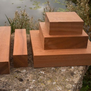 May include: Several wooden blocks of varying sizes and shapes, displayed on a stone surface. The blocks are a warm, reddish-brown hue, suggesting hardwood. The arrangement includes rectangular and square blocks, with a natural backdrop of water and greenery.