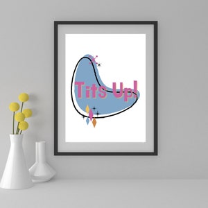 May include: A framed print with a blue, abstract shape and the text "Tits Up!" in pink letters. The background is white.
